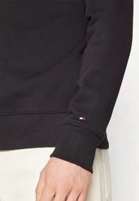 Persona che indossa una felpa nera a maniche lunghe con un piccolo logo rosso, bianco e blu sul polsino, abbinata a pantaloni color avorio.