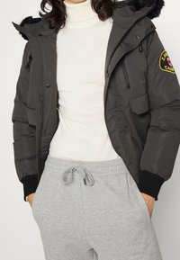 Puffervest i mørkegrå stof med sorte detaljer og hætte af kunstpels, parret med en cremet turtleneck og grå sweatpants.