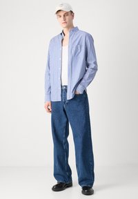 Koszula na guziki w paski w kolorach niebieskim i białym, noszona na białym topie, zestawiona z luźnymi niebieskimi dżinsami i czarnymi butami. Nieformalna czapka dopełnia stylizację.