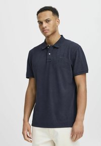 Tumši zilas krāsu polos krekls, kas izgatavots no mīksta auduma, ar klasisku apkakli, īsām piedurknēm un smalku izšūtu logotipu uz krūtīm.