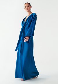 Lange blauwe satijnen jurk met een diepe V-hals, vloeiende mouwen en een gestrikte taille. Gladde textuur, volledige lengte met een lichte glans.