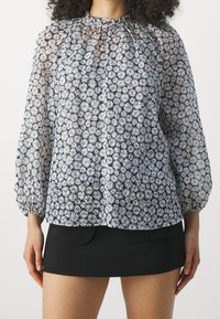 Blouse transparente avec encolure froncée, motif floral en bleu et blanc, manches longues bouffantes et coupe décontractée, associée à une mini jupe noire.