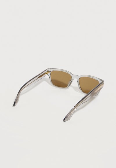 Γυαλιά ηλίου με διάφανο σκελετό και καφέ φακούς σε ορθογώνιο σχήμα, τοποθετημένα πάνω σε λευκή επιφάνεια.