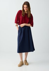 Femme debout, la tête légèrement baissée, portant un haut rouge ample, une jupe midi bleu marine avec des poches, des baskets beige et un bracelet au poignet gauche.