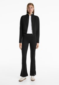 Crna zip-up krojena jakna s visokim ovratnikom, uparena s crnim širokim hlačama. Outfit ima glatku teksturu i minimalistički dizajn.