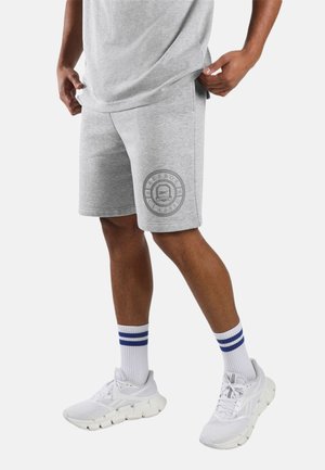 Man draagt lichtgrijze Reebok kortebroek met voorzakken, witte sneakers en witte sokken met twee blauwe strepen, staand op een witte achtergrond.