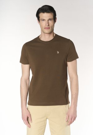 Uomo che indossa una maglietta marrone a maniche corte con un piccolo logo sul petto e pantaloni beige chiaro, in piedi davanti a uno sfondo chiaro uniforme.