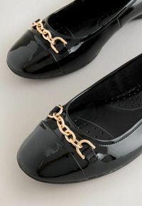 Ballerines en cuir verni noir avec une ceinture en chaîne dorée sur le cou-de-pied, dotées d'une texture lisse et d'un design à bout rond.