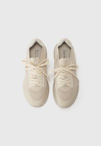 Beige sneakers with a breathable mesh upper, padded collar, and lace-up closure. Flat, textured sole with a subtle pattern.