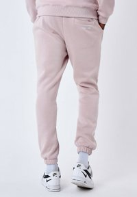 Pantalones de chándal suaves de color rosa pálido con puños de canalé, un bolsillo trasero y un pequeño logo blanco. Se llevan con zapatillas blancas y calcetines gris claro.