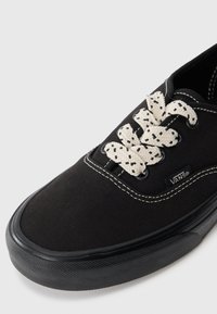 Crne Vans tenisice sa debelim bijelim vezicama koje sadrže crne mrlje, bijelim šavovima i teksturiranim crnim gumeni potplat.