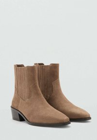 Botas de ante marrón hasta el tobillo con puntera puntiaguda, paneles laterales elásticos y un tacón bajo tipo bloque; textura suave y diseño minimalista.