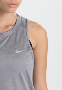 Primer plano de una persona que lleva una camiseta deportiva sin mangas gris de Nike con un sujetador deportivo negro debajo, sobre un fondo liso.