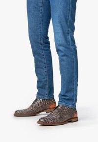 Piernas que llevan vaqueros azules y zapatos de cuero marrón oscuro con textura, de cordones y suela de madera, sobre un fondo blanco.