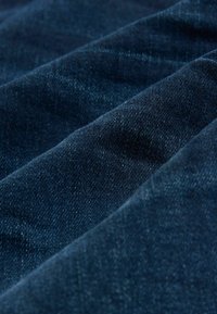 Tmavě modrá denímová látka s hladkou strukturou, projevující jemný lesk a mírné barevné odchylky, ukazující poskládané vrstvy a hloubku.