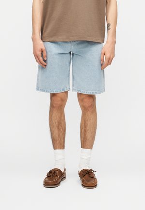 Hombre con pantalones cortos de mezclilla azul claro, zapatos casuales marrones con cordones, calcetines blancos y camiseta marrón, de pie contra un fondo liso.