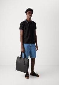 Černé polo triko s bílým lemováním límečku, džíny šortky, černá taška a sandály. Model stojí proti jednoduchému pozadí.