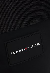 Fekete szövet, textúrált felszínnel, fekete téglalap alakú logópacsival, amelyen fehér szöveg olvasható: "TOMMY HILFIGER", valamint színes fókuszpontok.