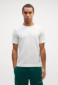 Svetlošedé bavlnené tričko s krátkymi rukávmi, okrúhlym výstrihom a modrým logom "Calvin Klein" na ľavej strane. Nosené s zelenými nohavicami.