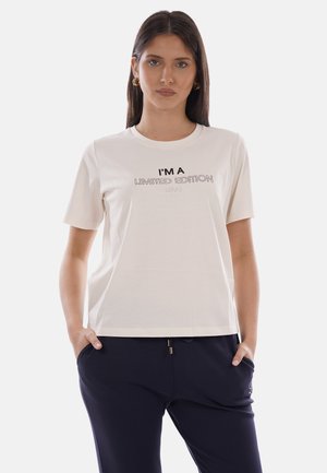 Donna che indossa una maglietta bianca con la scritta "I'M A LIMITED EDITION", pantaloni blu navy, in piedi con le mani nelle tasche davanti a uno sfondo semplice.