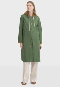 Imperméable vert à capuche, avec une fermeture à brandebourg beige et des cordons de serrage. Tissu texturé, longueur jusqu'aux genoux, avec des poches latérales.