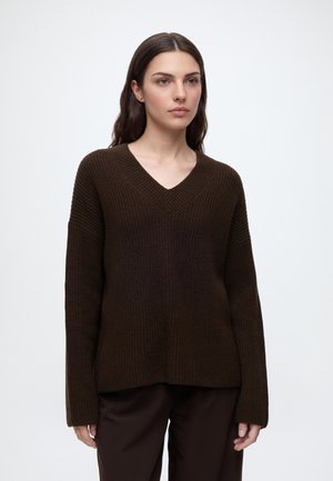 Femme aux longs cheveux bruns portant un pull en maille brun foncé à col en V et un pantalon foncé, debout devant un fond clair uni.