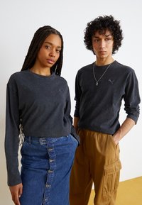 Două persoane poartă bluze negre cu mâneci lungi, cu o textură subtilă și un logo. Una este îmbrăcată cu o fustă albastră cu nasturi, iar cealaltă poartă pantaloni cargo bej.