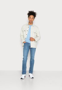 Chaqueta de botón gris claro con acentos en los bolsillos negros, superpuesta a una camiseta azul clara, combinada con jeans azules y zapatillas blancas.
