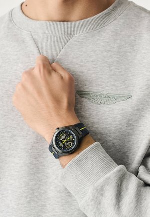 Férfi világosszürke Aston Martin pulóverben, keze a nyakánál, bal csuklóján fekete és sárga sportóra látható.