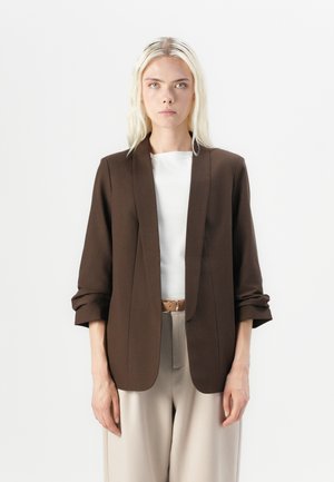 Ruskea bleiseri, jossa on muotoiltu istuvuus, notched-lapels ja käännetyt hihat, yhdistetty valkoiseen puseroon ja beigeihin housuihin.