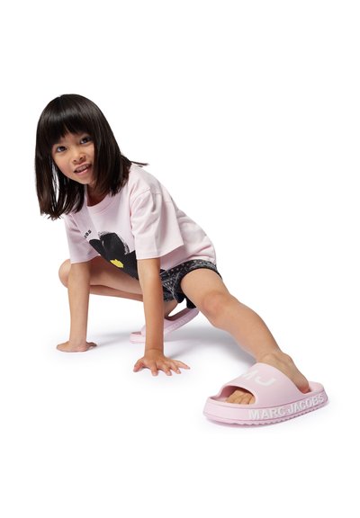 Infradito in gomma rosa con una fascia larga che presenta la scritta "MARC JACOBS" in caratteri bianchi. L'outfit del bambino include una t-shirt rosa chiaro e pantaloni corti neri.