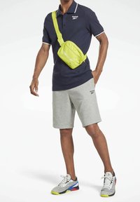 Reebok polo tričko v námořnické modři s bílým rámečkem, šedé šortky a jasně zelená crossbody taška. Tenisky mají prodyšný design s červenými, modrými a žlutými akcenty.