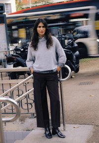 Pulover gri deschis cu guler înalt, pantaloni largi maro închis și cizme negre lucioase. Balustradă metalică și autobuze neclare în fundal.