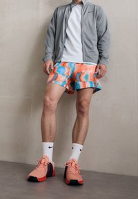 Sportsoutfit bestående af en grå lynlåsjakke, hvid T-shirt, farverige shorts med abstrakt print og pink løbesko med sorte detaljer.