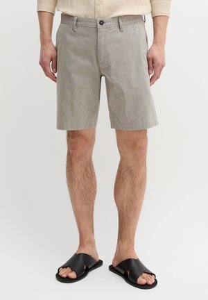 Man draagt lichtgrijze korte broek, beige overhemd en zwarte sandalen met gekruiste bandjes, staand tegen een effen achtergrond.