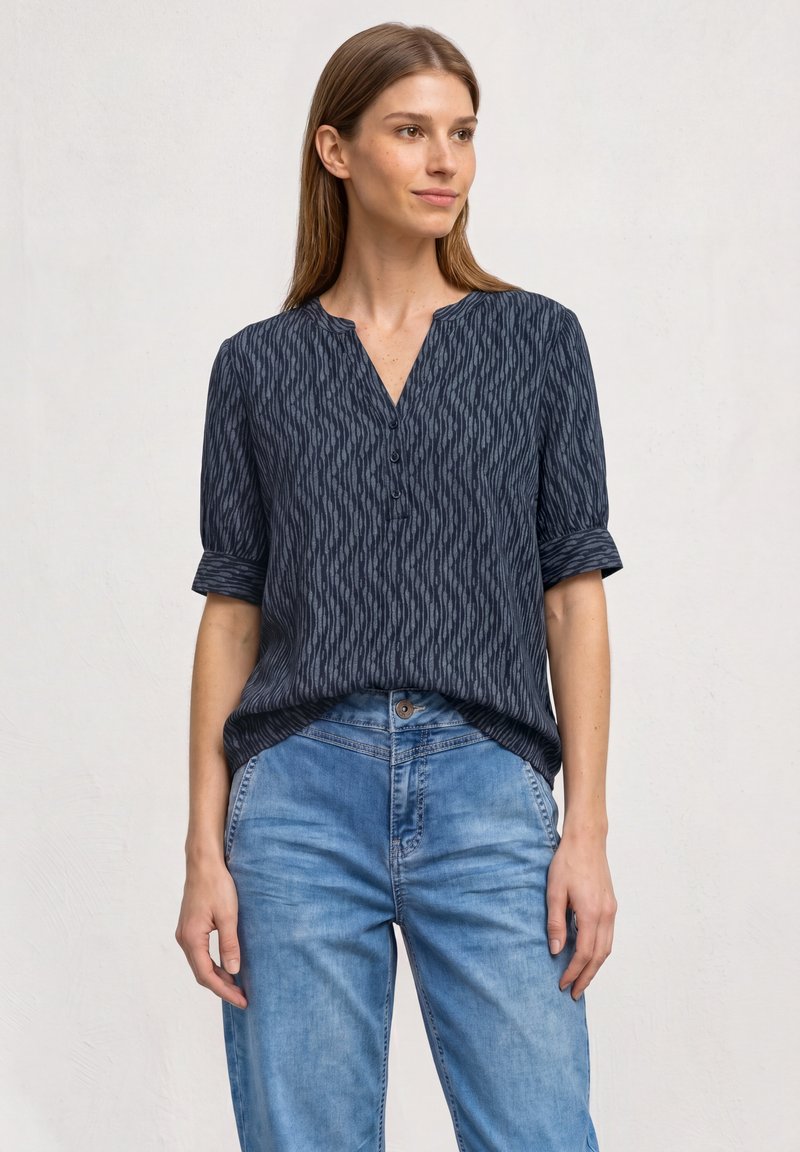 Femme portant un chemisier foncé à motifs boutonné avec les manches retroussées et un jean bleu clair, debout devant un fond gris clair uni.