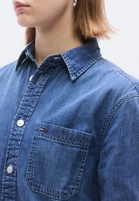 Jeans srajca v srednje modri barvi, z ostrim ovratnikom, zapiranjem na gumbe in levim žepom na prsih. Tkanina z teksturo in vidnimi šivi.