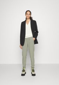 Blazer preto sobre um suéter de malha branco com decote em V, combinado com calças verde-claro e botas de amarrar bege com solas pretas e detalhes verdes.
