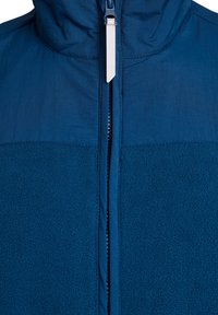 Granatowa kurtka rozpinana z połączenia gładkiego nylonu i fakturowanej polarowej tkaniny. Posiada stojący kołnierz i centralny zamek błyskawiczny.