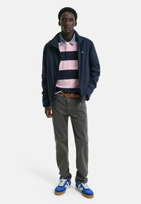 Giacca leggera navy sopra una polo a righe rosa e navy, pantaloni grigi e sneakers blu con accenti bianchi e cintura marrone.