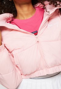 Kvinna iklädd en ljus rosa pufferjacka som är öppen över en rosa skjorta och en vit plisserad kjol.
