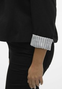 Haut noir à manches longues avec un poignet contrastant blanc à fines rayures verticales, associé à un pantalon noir ajusté. Texture de tissu lisse.