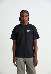 Black cotton t-shirt with short sleeves. Features a small pink logo on the left chest. Accompanied by grey pants, standing against a plain background.