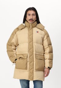 Beige dunjakke med kviltet design, høy krage, frontlommer og logo-montert. Har en glatt tekstur og kontrasterende fargeblokker.