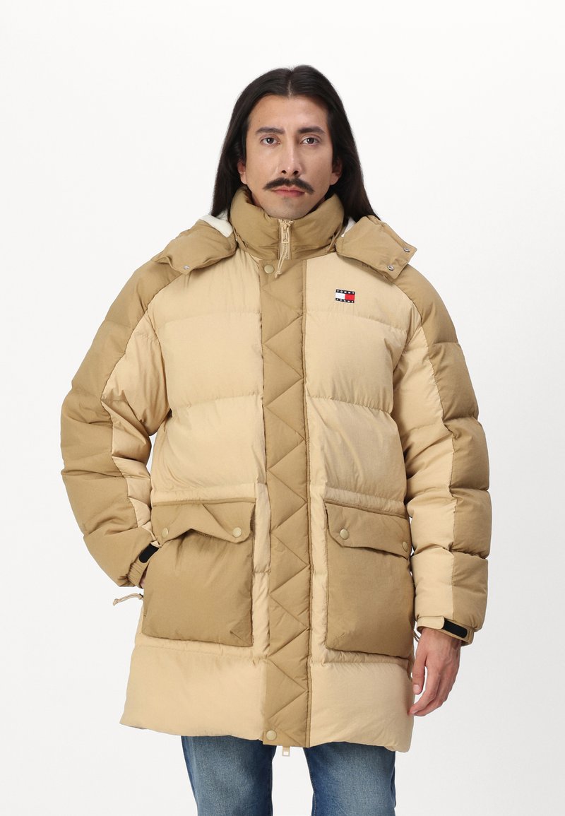 Beige dunjakke med kviltet design, høy krage, frontlommer og logo-montert. Har en glatt tekstur og kontrasterende fargeblokker.