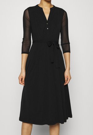 Schwarzes Midikleid mit transparenten Dreiviertelärmeln, V-Ausschnitt mit Knöpfen und einem gebundenen Taillengürtel an einer stehenden Person.