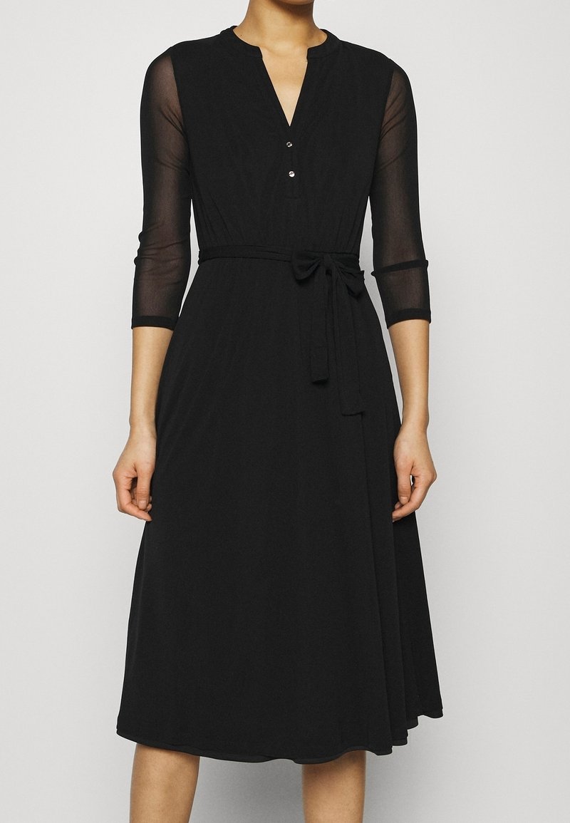 Robe midi noire avec manches transparentes trois-quarts, col en V avec boutons, et ceinture nouée à la taille sur une personne debout.