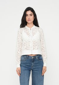 Blouson aviateur en dentelle blanche avec fermeture éclair devant, présentant un motif floral, des poignets côtelés et une coupe courte, porté avec un jean taille haute.