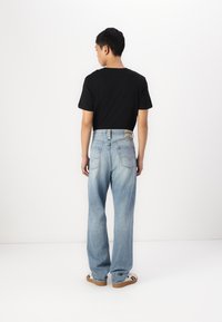 Jeans de mezclilla de pierna ancha en azul claro con un parche en la cinturilla trasera, combinados con una camiseta negra y zapatillas sobre un fondo liso.