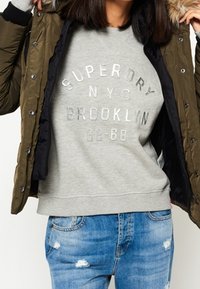 Chaqueta acolchada verde oliva con capucha de piel sintética, combinada con una sudadera gris clara con texto plateado y vaqueros azules desgastados.